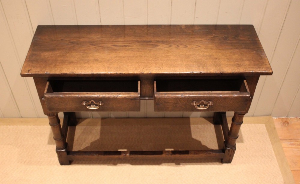 A solid oak server table featuring a rectangular top, two drawers with brass swan-neck handles, turned front legs, and a lower stretcher base. View VI