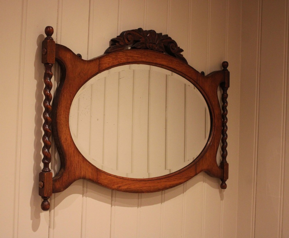 An oval glass wall mirror set within a dark carved oak frame featuring turned barley twist side pilasters and an ornate cresting.