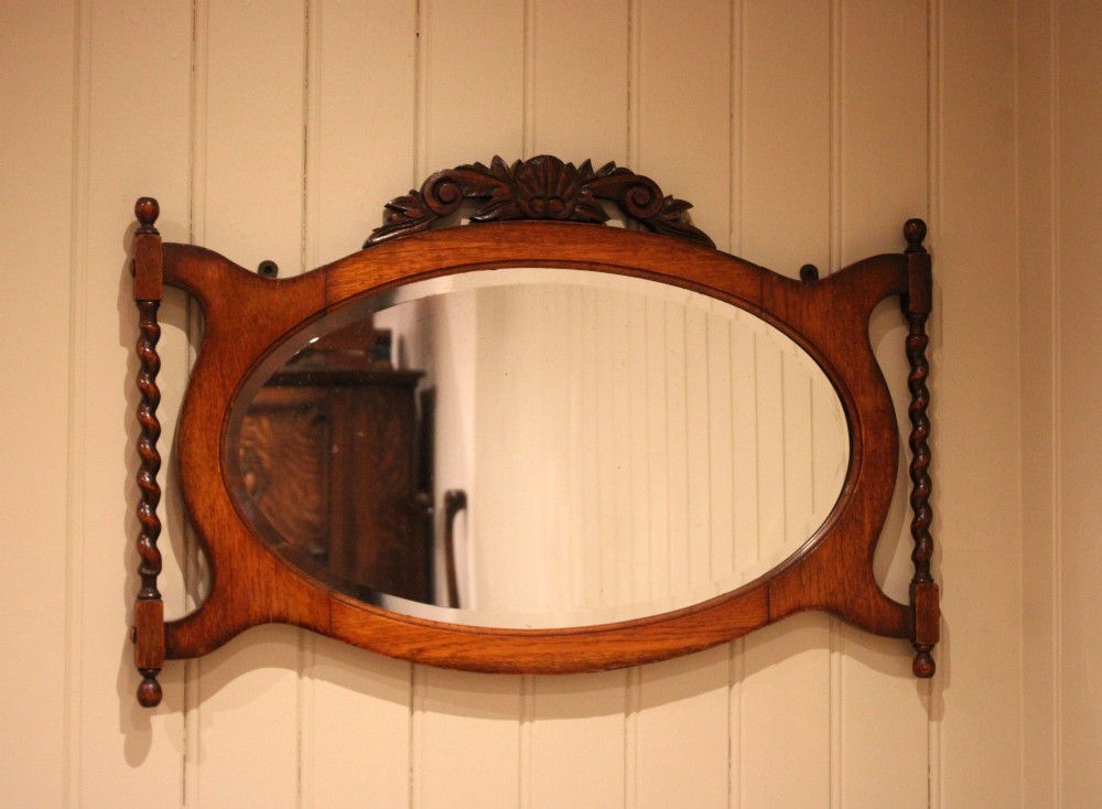 An oval glass wall mirror set within a dark carved oak frame featuring turned barley twist side pilasters and an ornate cresting. View II