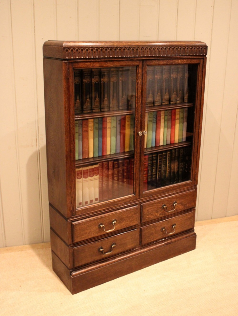 A medium brown oak glazed bookcase with two glass upper doors, carved egg-and-dart top moulding, and four drawers with brass handles.