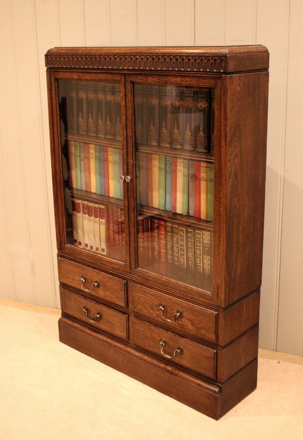 A medium brown oak glazed bookcase with two glass upper doors, carved egg-and-dart top moulding, and four drawers with brass handles. View V