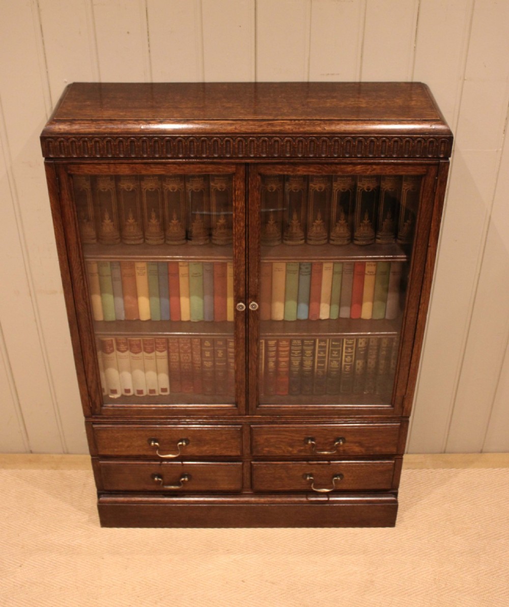 A medium brown oak glazed bookcase with two glass upper doors, carved egg-and-dart top moulding, and four drawers with brass handles. View VII