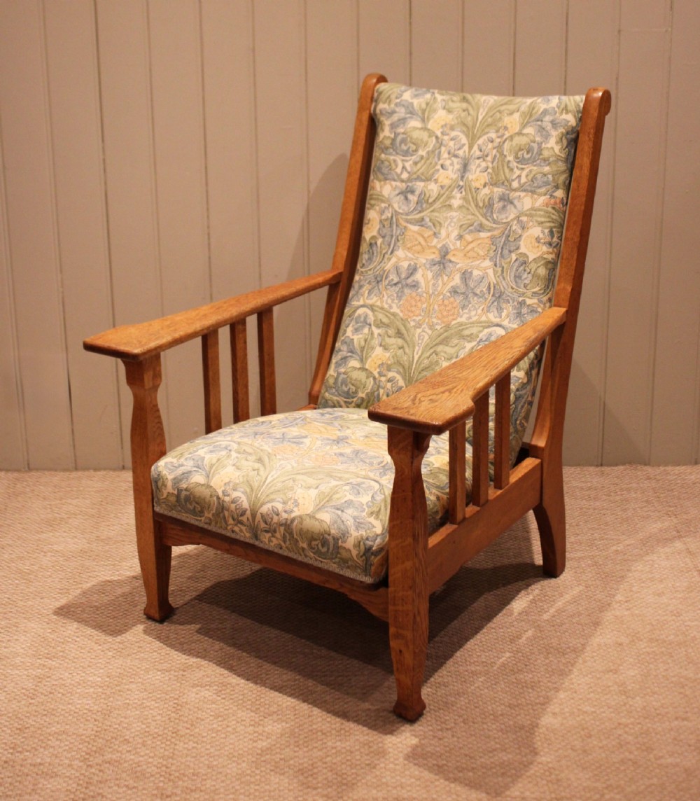 A solid oak Arts and Crafts style armchair featuring broad flat armrests and upholstery decorated with a botanical foliage pattern.