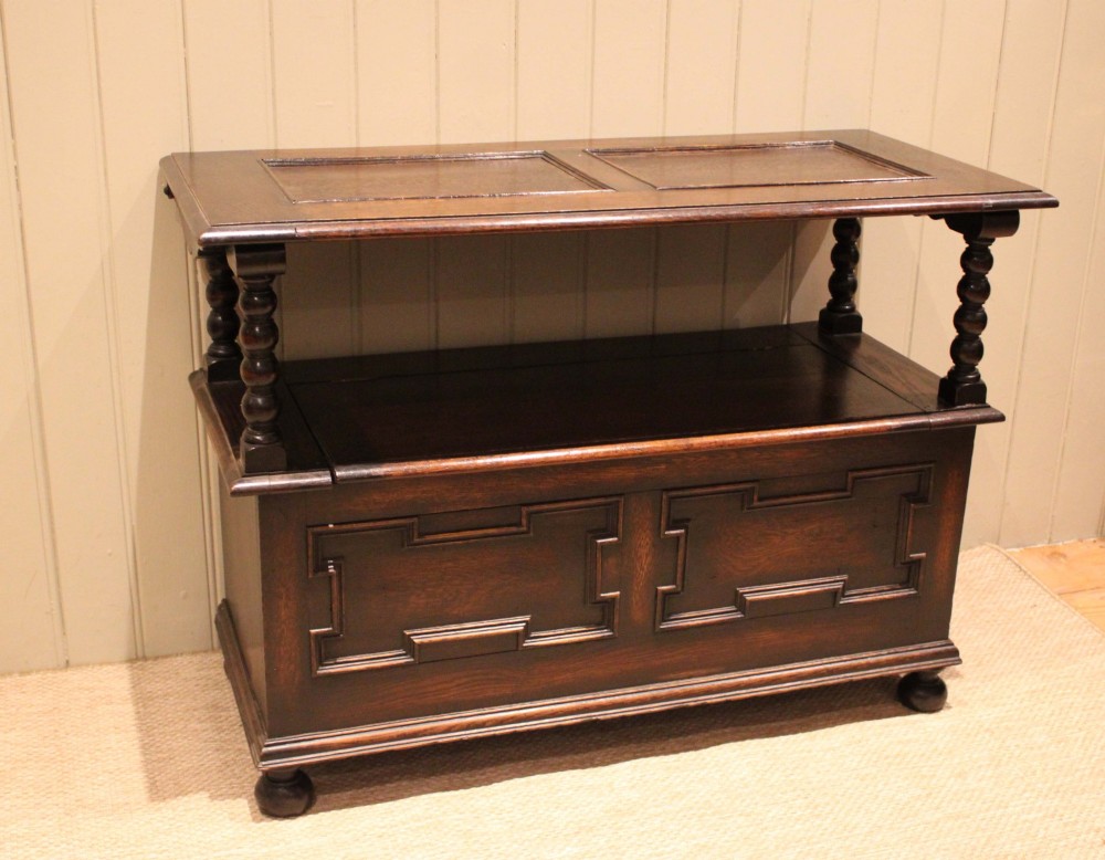 A dark oak monks bench with geometric moulded panels on the backrest and storage base, supported by turned posts and bun feet. View II