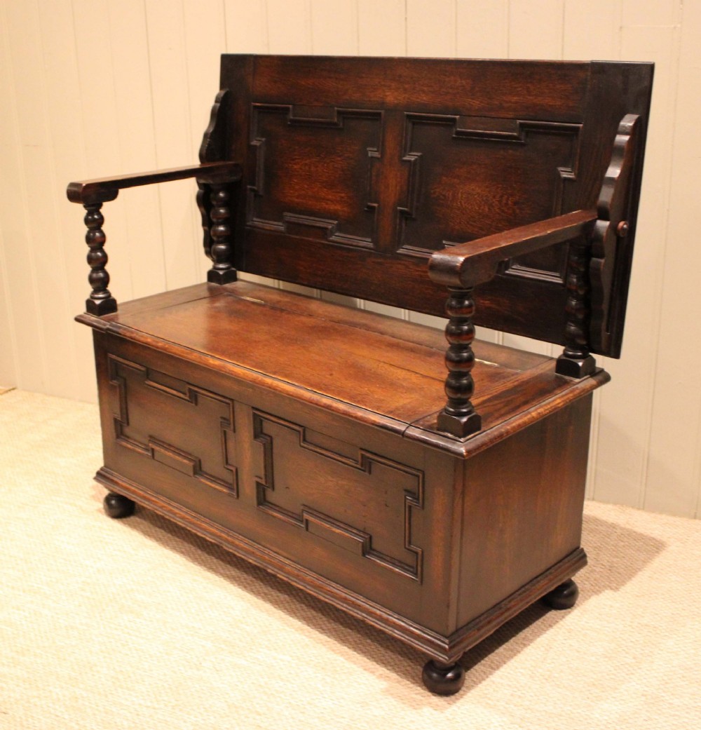 A dark oak monks bench with geometric moulded panels on the backrest and storage base, supported by turned posts and bun feet. View III