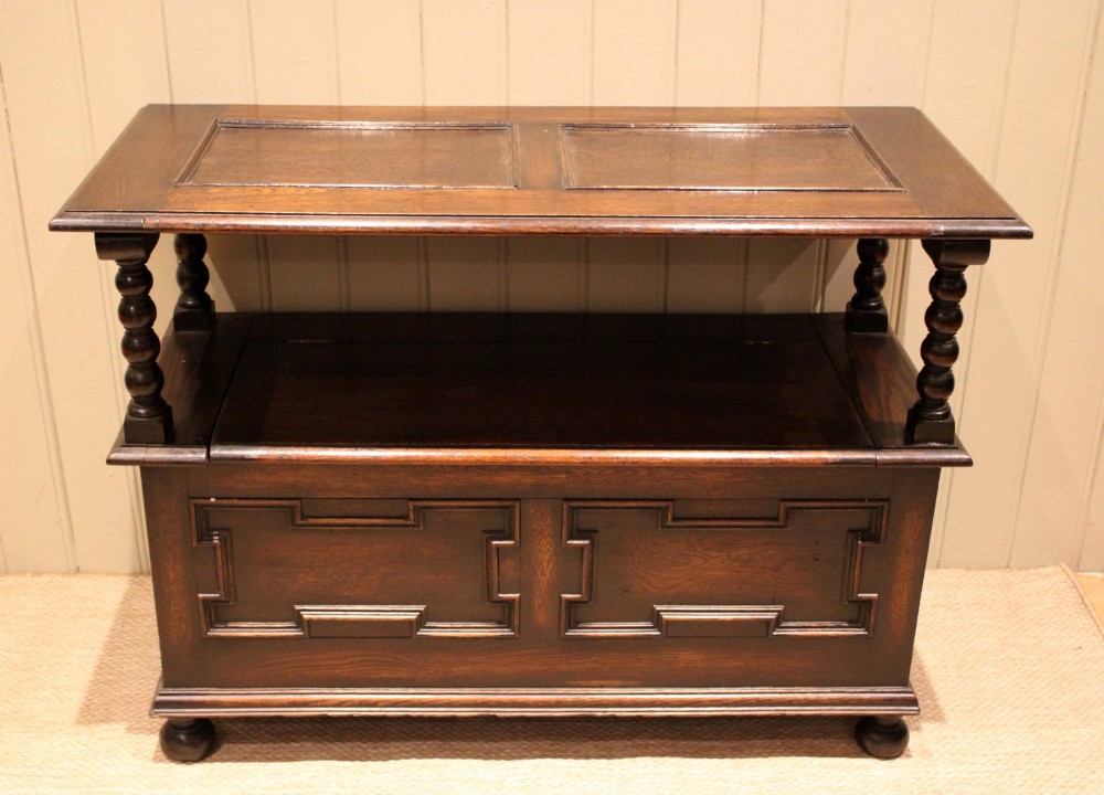 A dark oak monks bench with geometric moulded panels on the backrest and storage base, supported by turned posts and bun feet. View IV