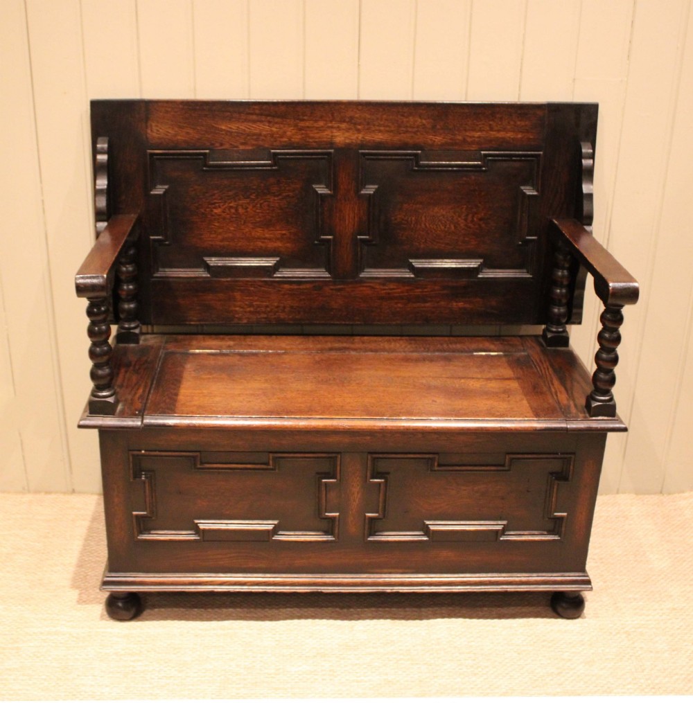 A dark oak monks bench with geometric moulded panels on the backrest and storage base, supported by turned posts and bun feet. View V