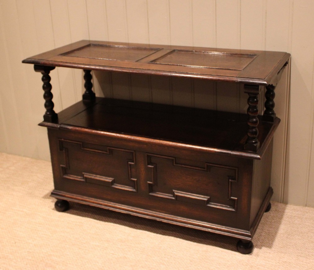 A dark oak monks bench with geometric moulded panels on the backrest and storage base, supported by turned posts and bun feet. View VI