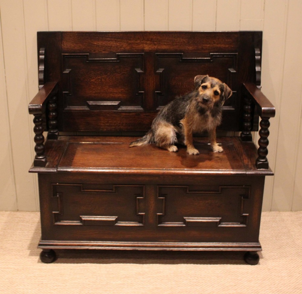 A dark oak monks bench with geometric moulded panels on the backrest and storage base, supported by turned posts and bun feet. View VII