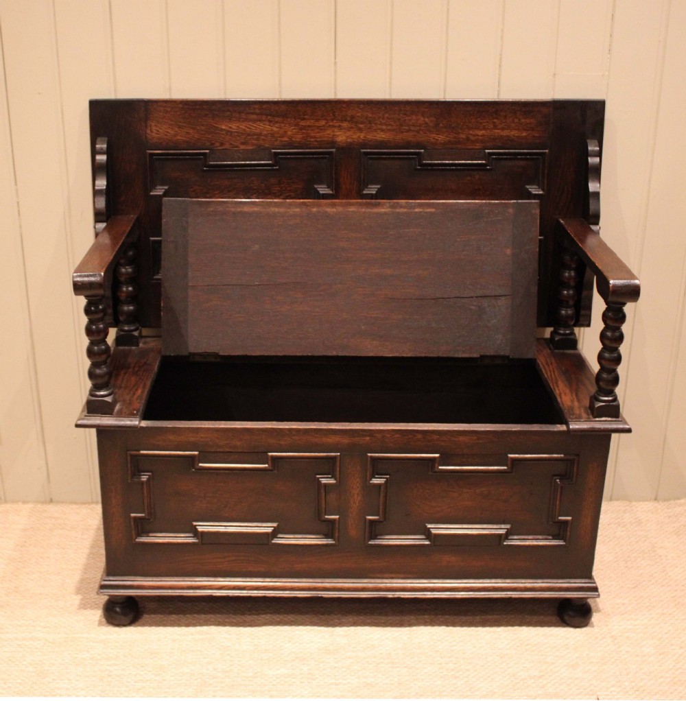 A dark oak monks bench with geometric moulded panels on the backrest and storage base, supported by turned posts and bun feet. View IX