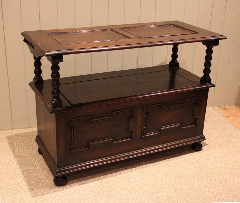 A dark oak monks bench with geometric moulded panels on the backrest and storage base, supported by turned posts and bun feet. View X