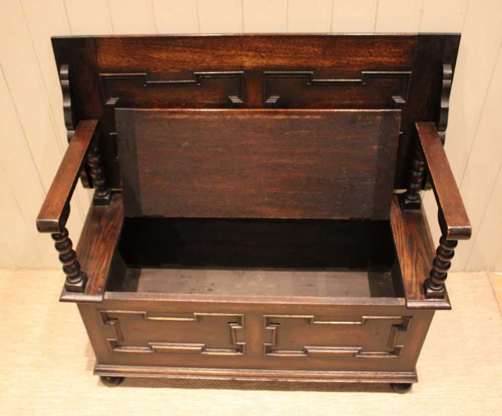 A dark oak monks bench with geometric moulded panels on the backrest and storage base, supported by turned posts and bun feet. View XI