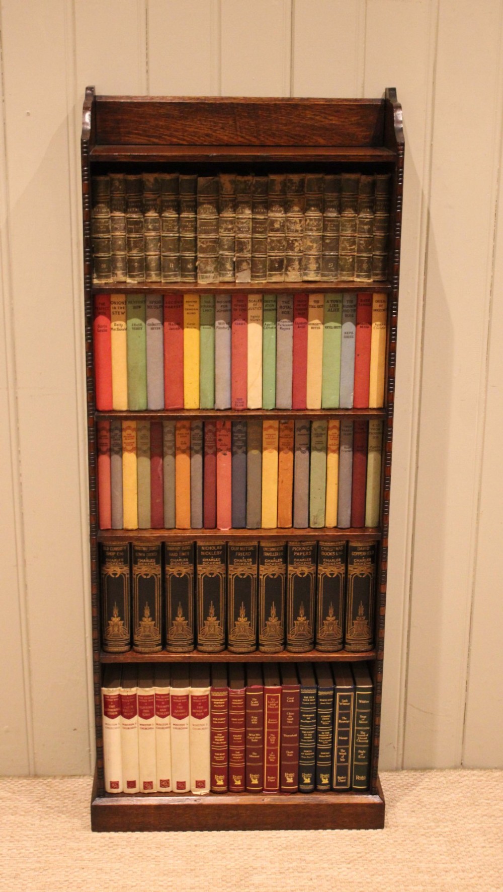 A tall, narrow dark oak open bookcase featuring five fixed shelves filled with vintage hardback books and a simple gallery top on a plinth base. View III