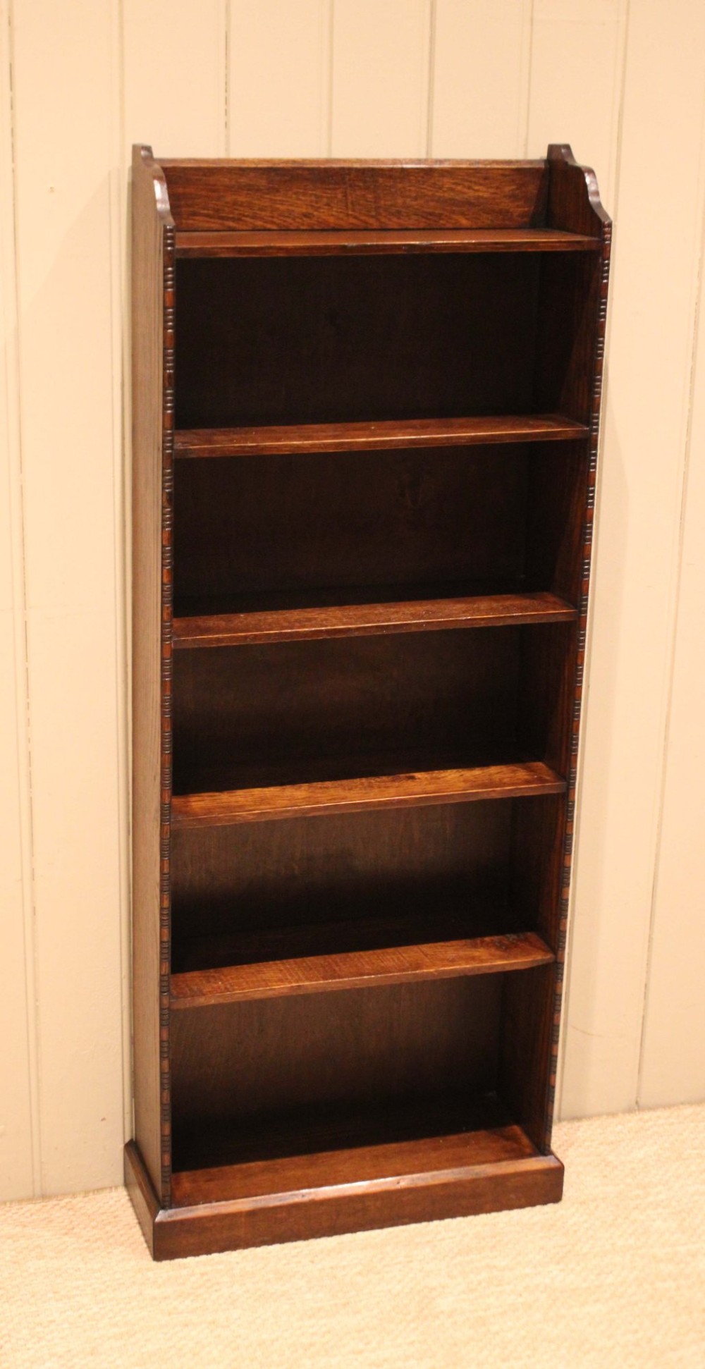 A tall, narrow dark oak open bookcase featuring five fixed shelves filled with vintage hardback books and a simple gallery top on a plinth base. View IV