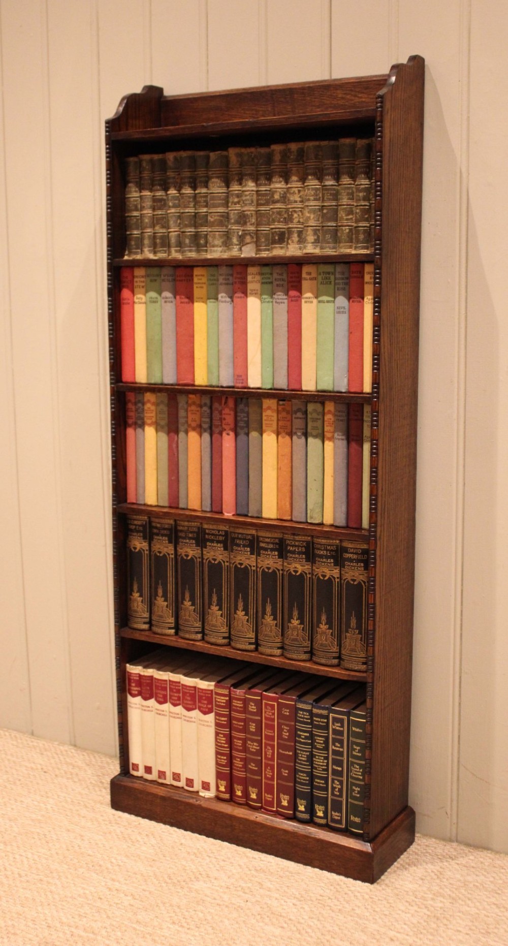 A tall, narrow dark oak open bookcase featuring five fixed shelves filled with vintage hardback books and a simple gallery top on a plinth base. View V