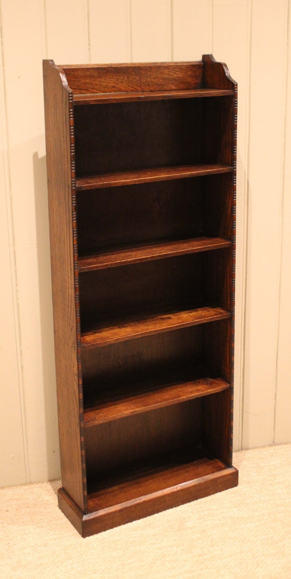 A tall, narrow dark oak open bookcase featuring five fixed shelves filled with vintage hardback books and a simple gallery top on a plinth base. View VI