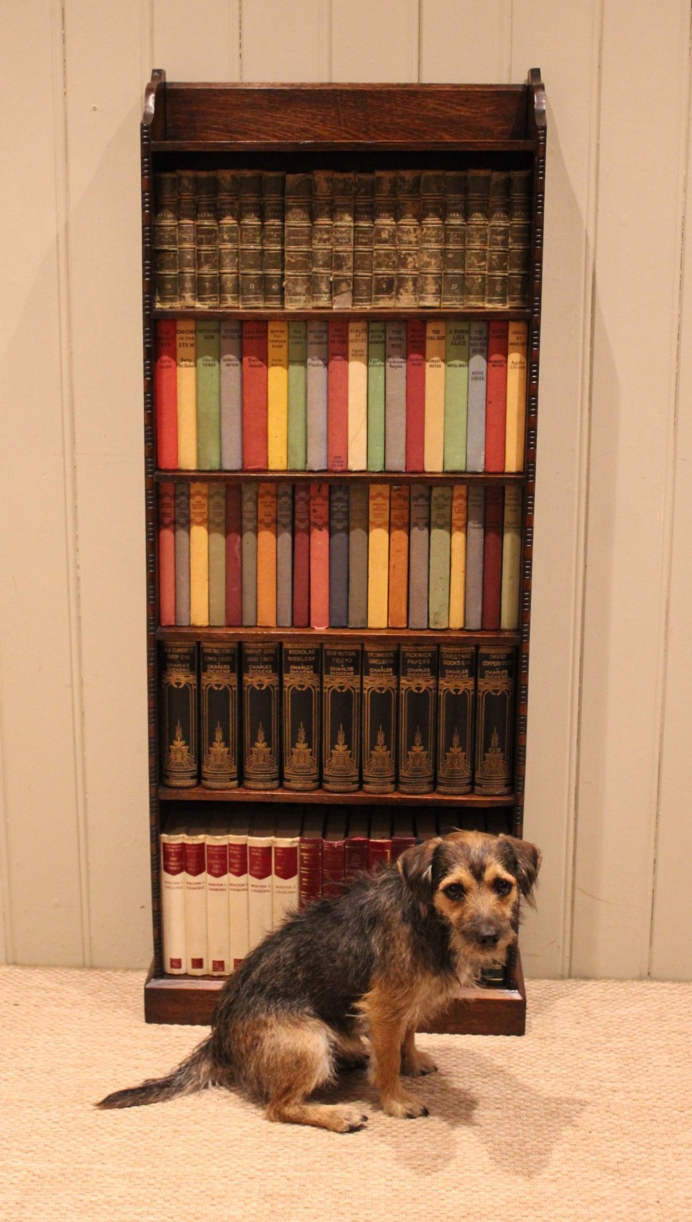 A tall, narrow dark oak open bookcase featuring five fixed shelves filled with vintage hardback books and a simple gallery top on a plinth base. View IX