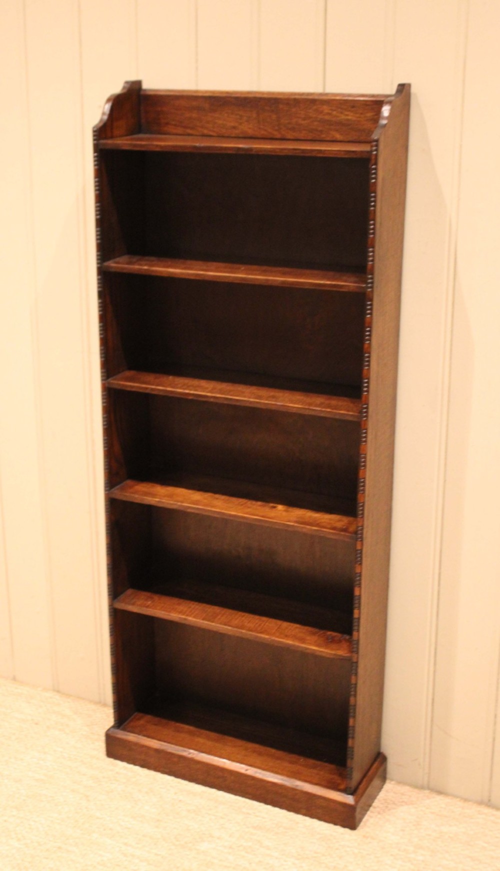 A tall, narrow dark oak open bookcase featuring five fixed shelves filled with vintage hardback books and a simple gallery top on a plinth base. View X