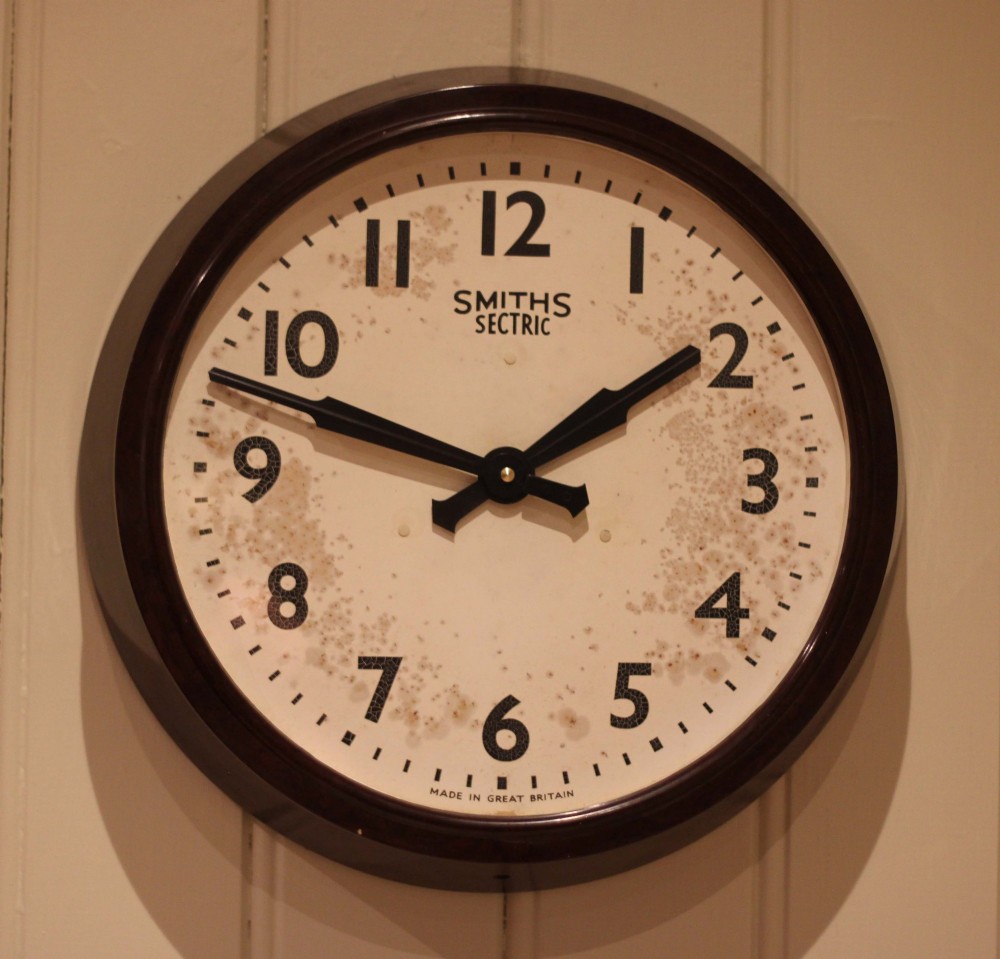 A circular vintage British Smiths Sectric wall clock featuring a dark brown Bakelite frame and an off-white dial with black Arabic numerals. View V