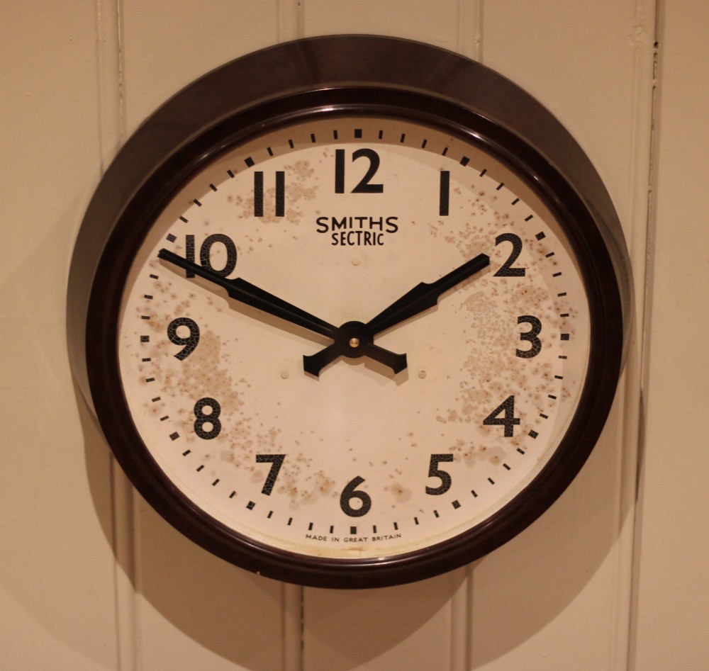 A circular vintage British Smiths Sectric wall clock featuring a dark brown Bakelite frame and an off-white dial with black Arabic numerals. View VII