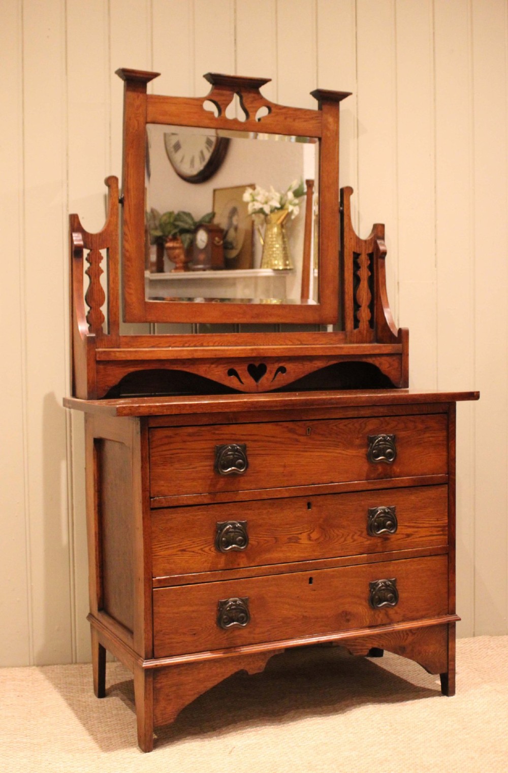 A solid oak Arts and Crafts dressing chest featuring three wide drawers, metal drop handles, heart-shaped cutouts, and a tilting rectangular mirror on top.