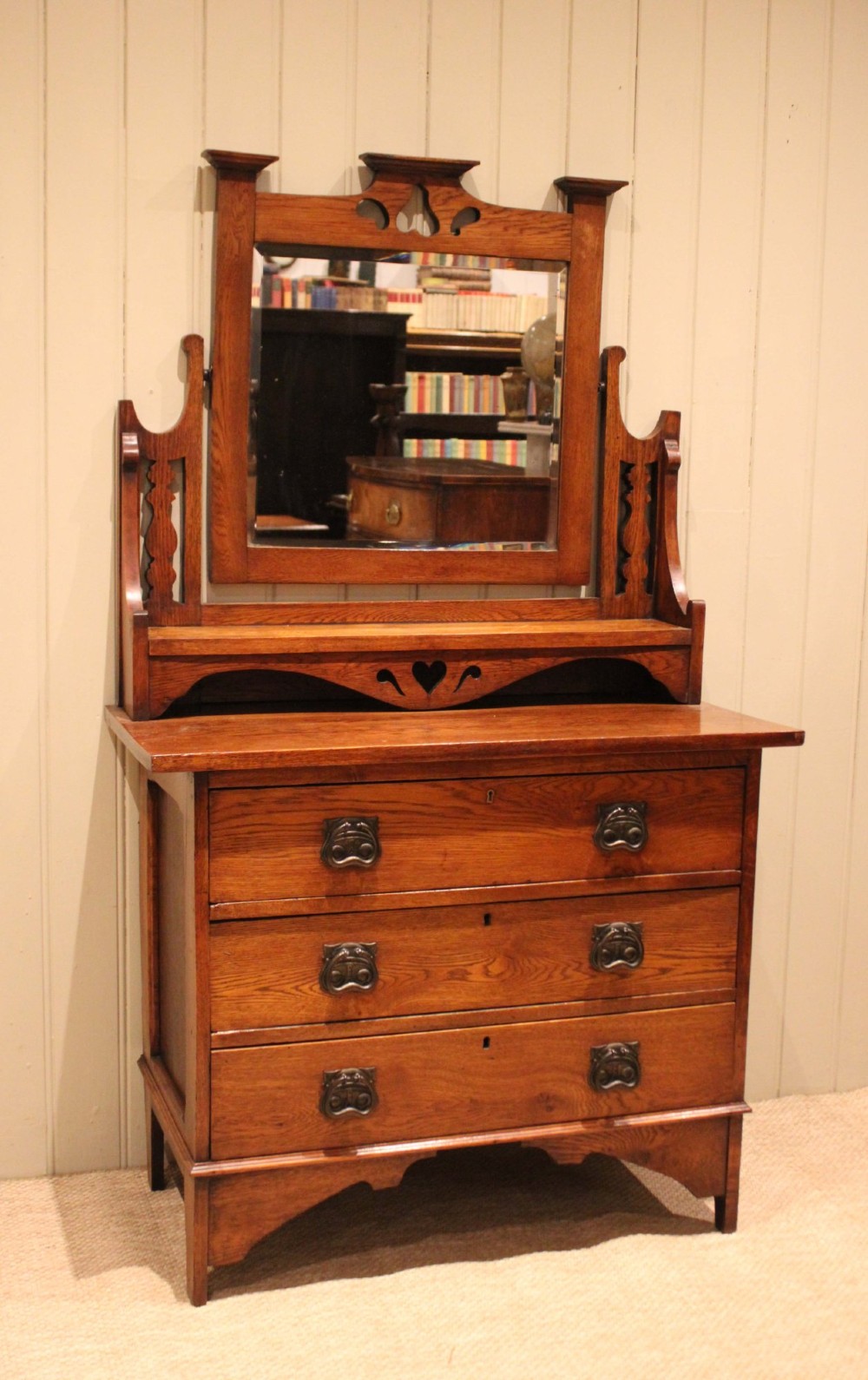 A solid oak Arts and Crafts dressing chest featuring three wide drawers, metal drop handles, heart-shaped cutouts, and a tilting rectangular mirror on top. View II