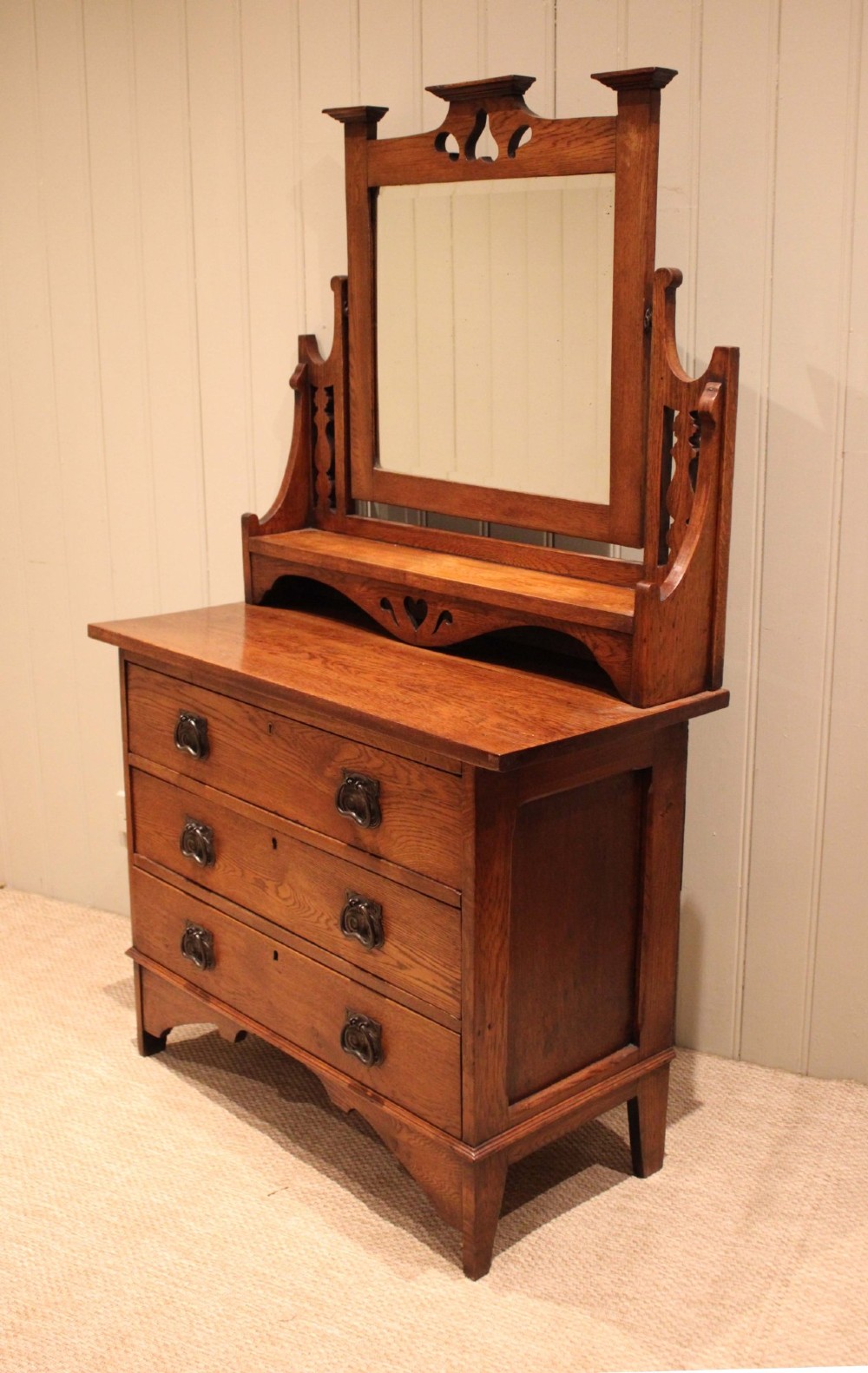 A solid oak Arts and Crafts dressing chest featuring three wide drawers, metal drop handles, heart-shaped cutouts, and a tilting rectangular mirror on top. View III