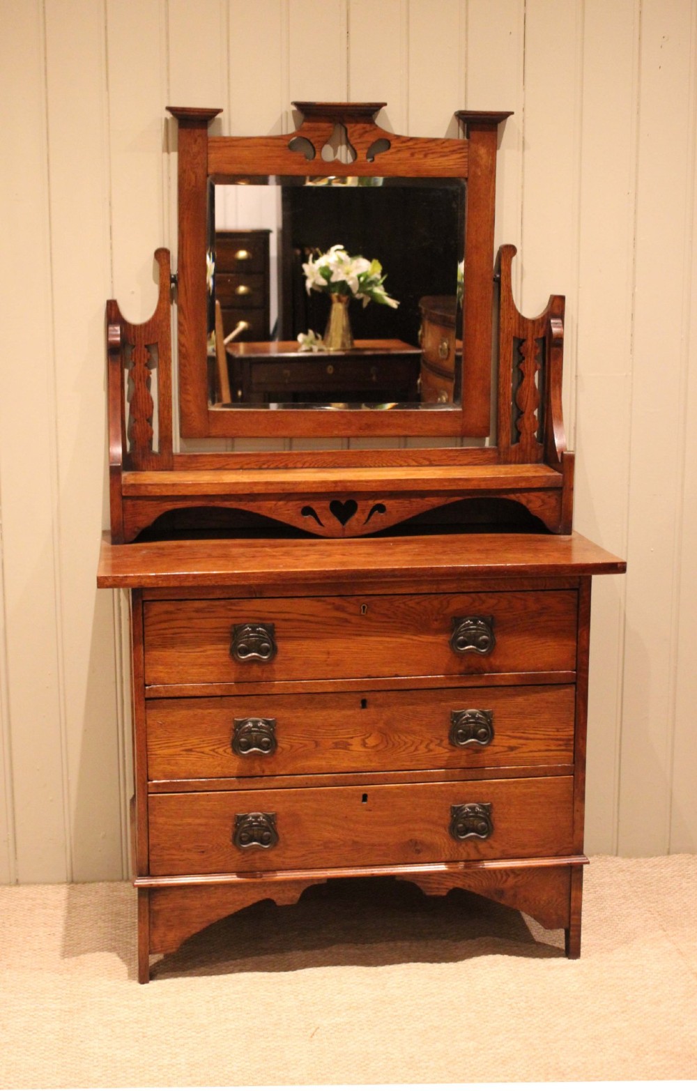 A solid oak Arts and Crafts dressing chest featuring three wide drawers, metal drop handles, heart-shaped cutouts, and a tilting rectangular mirror on top. View IV