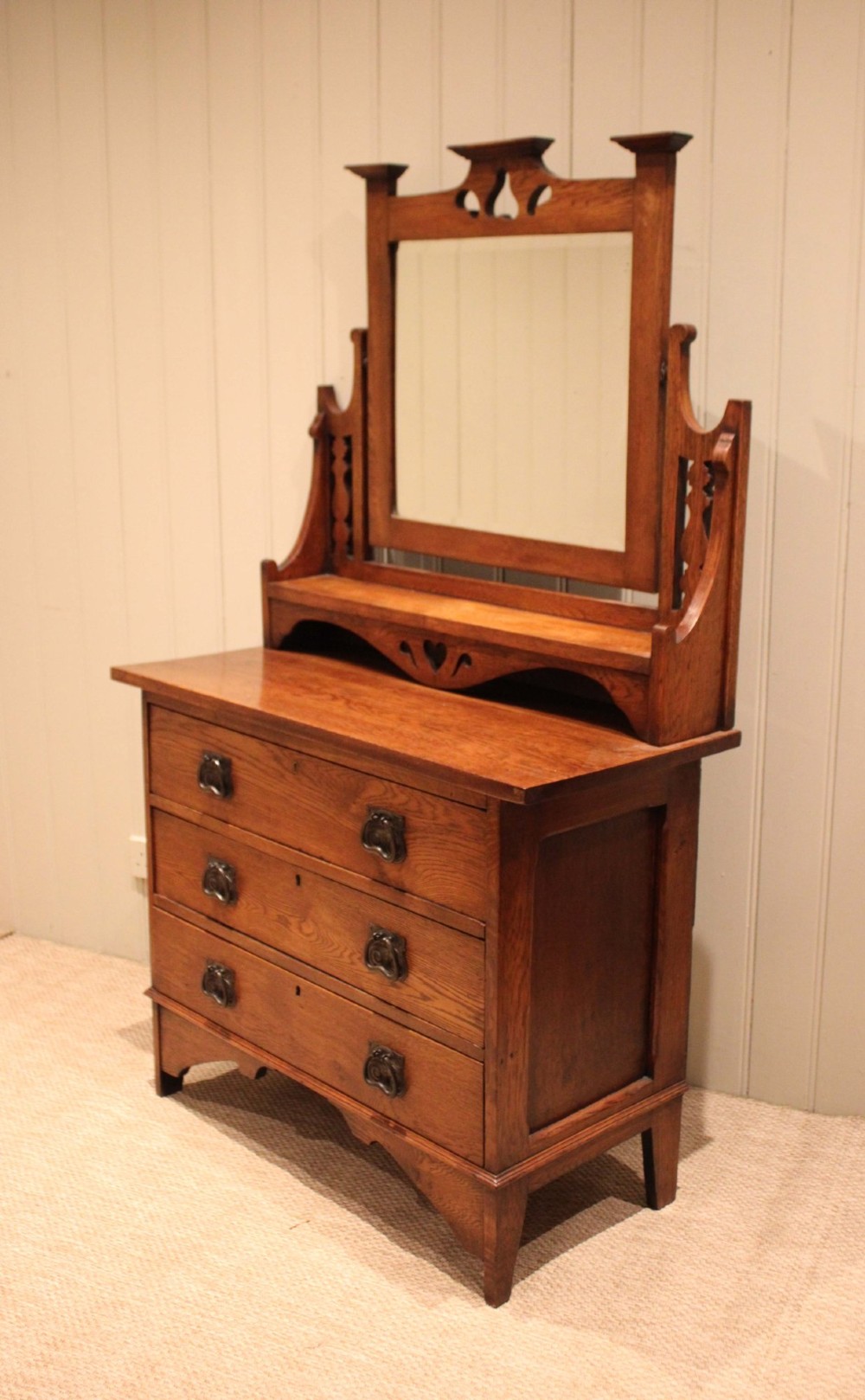 A solid oak Arts and Crafts dressing chest featuring three wide drawers, metal drop handles, heart-shaped cutouts, and a tilting rectangular mirror on top. View V