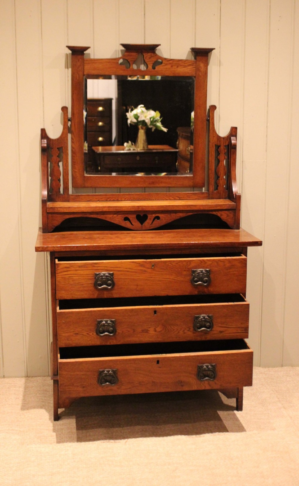 A solid oak Arts and Crafts dressing chest featuring three wide drawers, metal drop handles, heart-shaped cutouts, and a tilting rectangular mirror on top. View VI