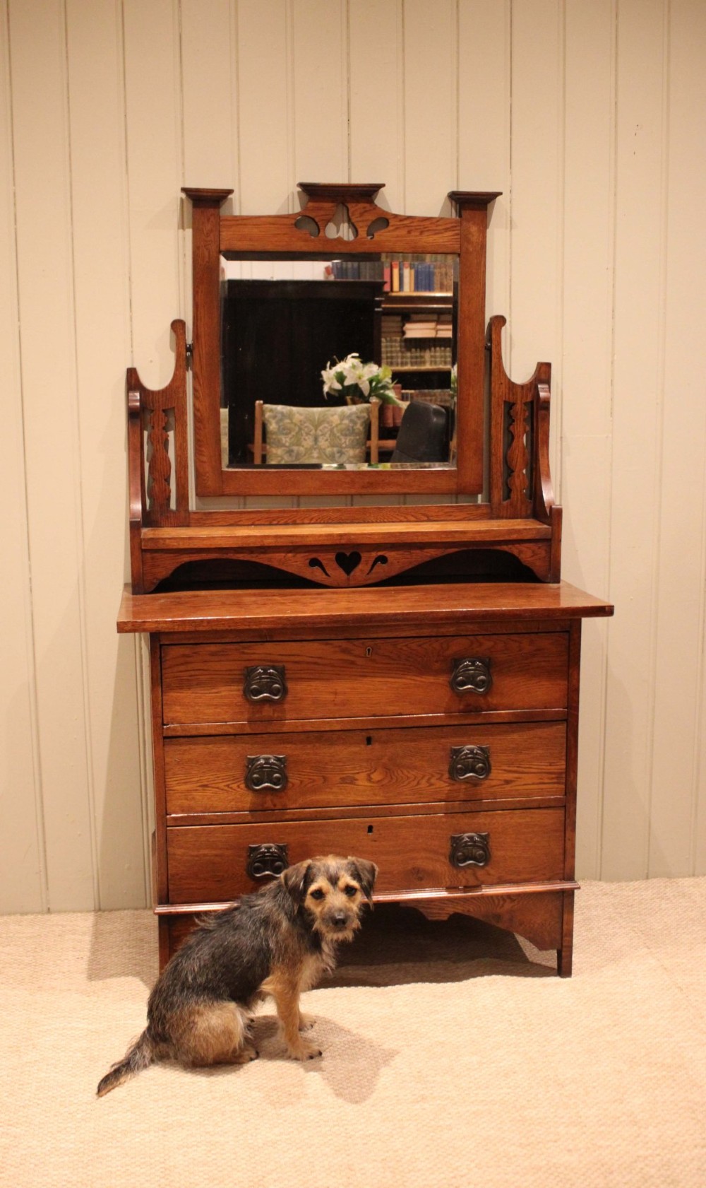 A solid oak Arts and Crafts dressing chest featuring three wide drawers, metal drop handles, heart-shaped cutouts, and a tilting rectangular mirror on top. View X