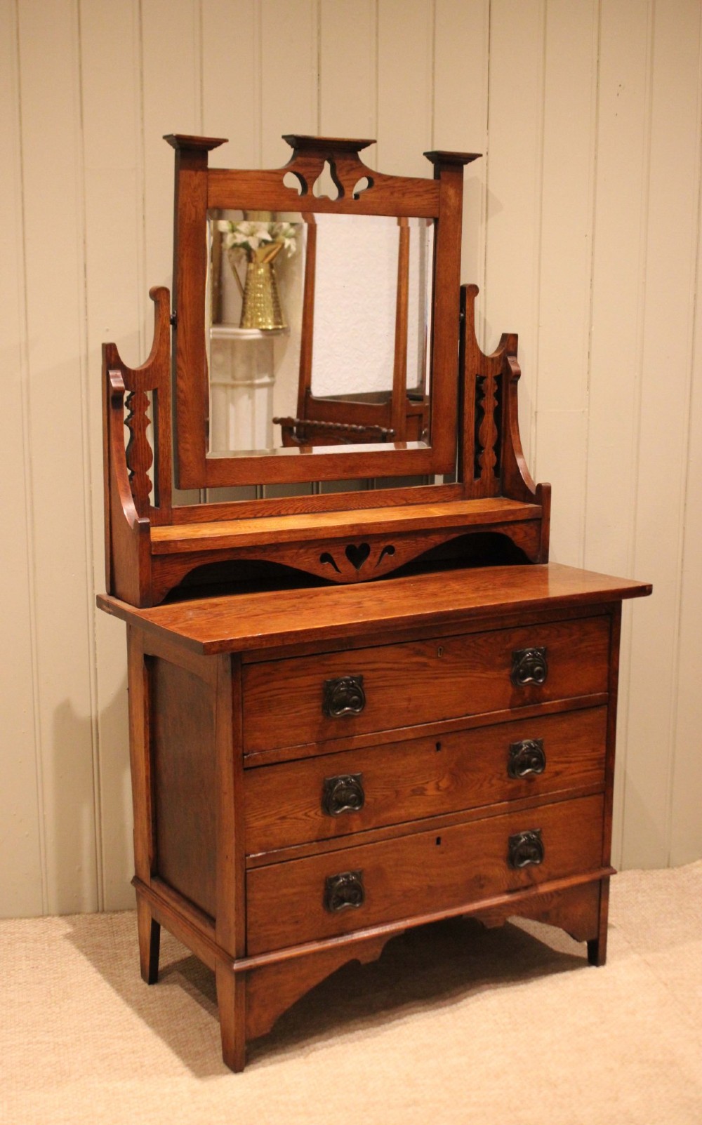 A solid oak Arts and Crafts dressing chest featuring three wide drawers, metal drop handles, heart-shaped cutouts, and a tilting rectangular mirror on top. View XI