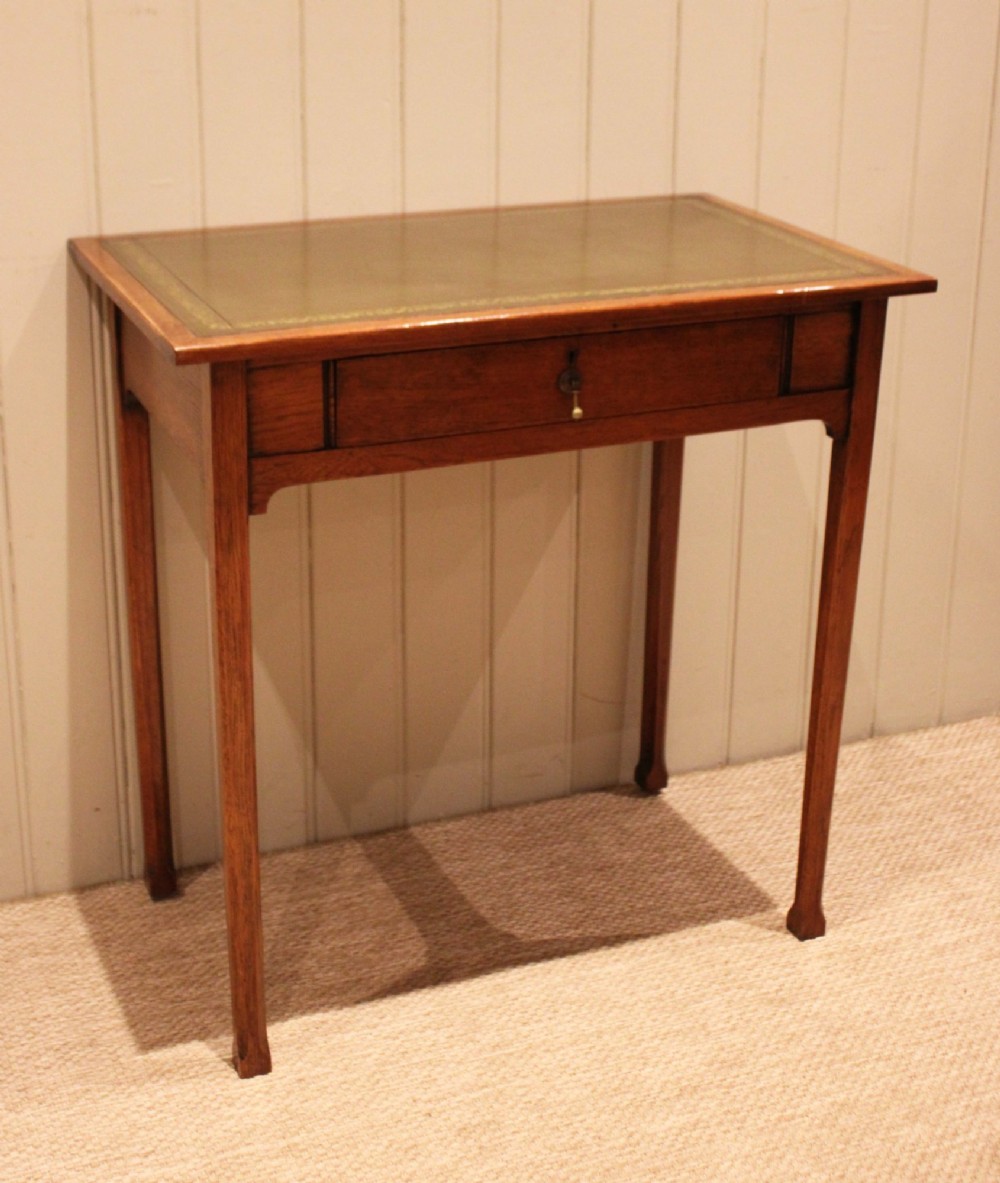 A small rectangular Arts and Crafts oak writing desk with a green gilded leather inset top, a single frieze drawer, and tapered square legs. View VII