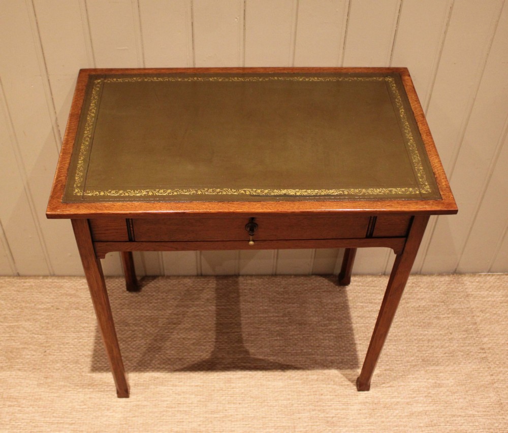 A small rectangular Arts and Crafts oak writing desk with a green gilded leather inset top, a single frieze drawer, and tapered square legs. View X