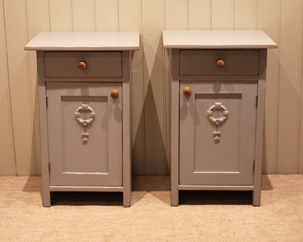 A pair of light grey painted pine bedside cabinets, each featuring a single upper drawer above a cupboard door decorated with a carved wreath motif.