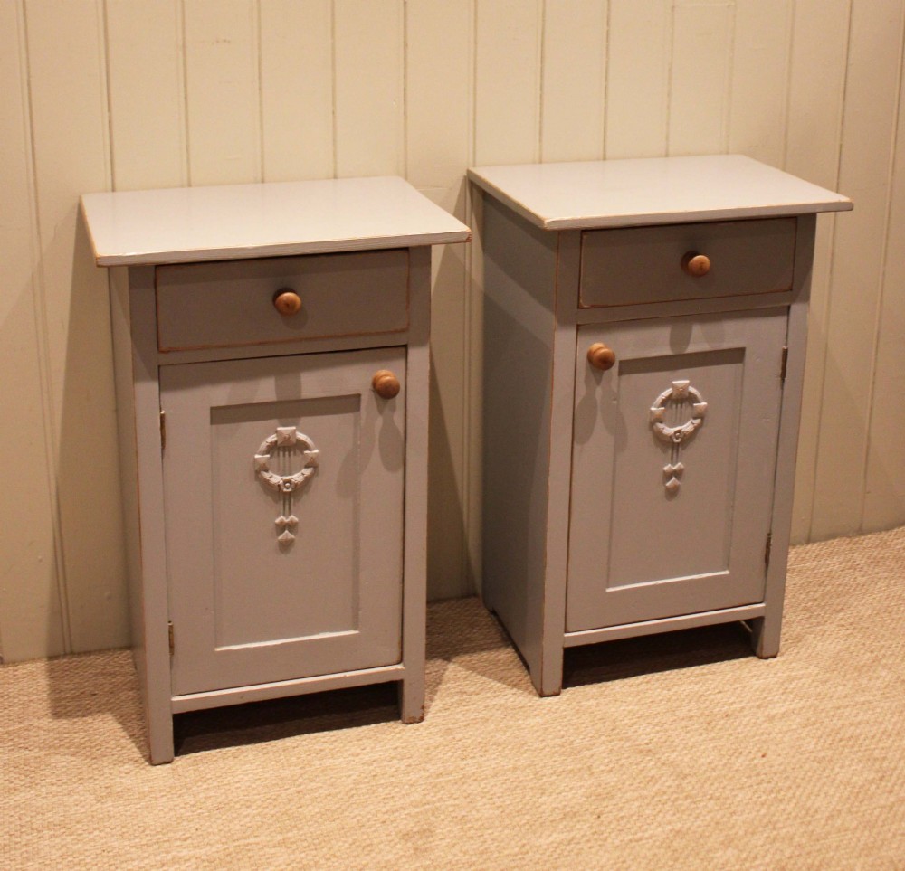 A pair of light grey painted pine bedside cabinets, each featuring a single upper drawer above a cupboard door decorated with a carved wreath motif. View IV