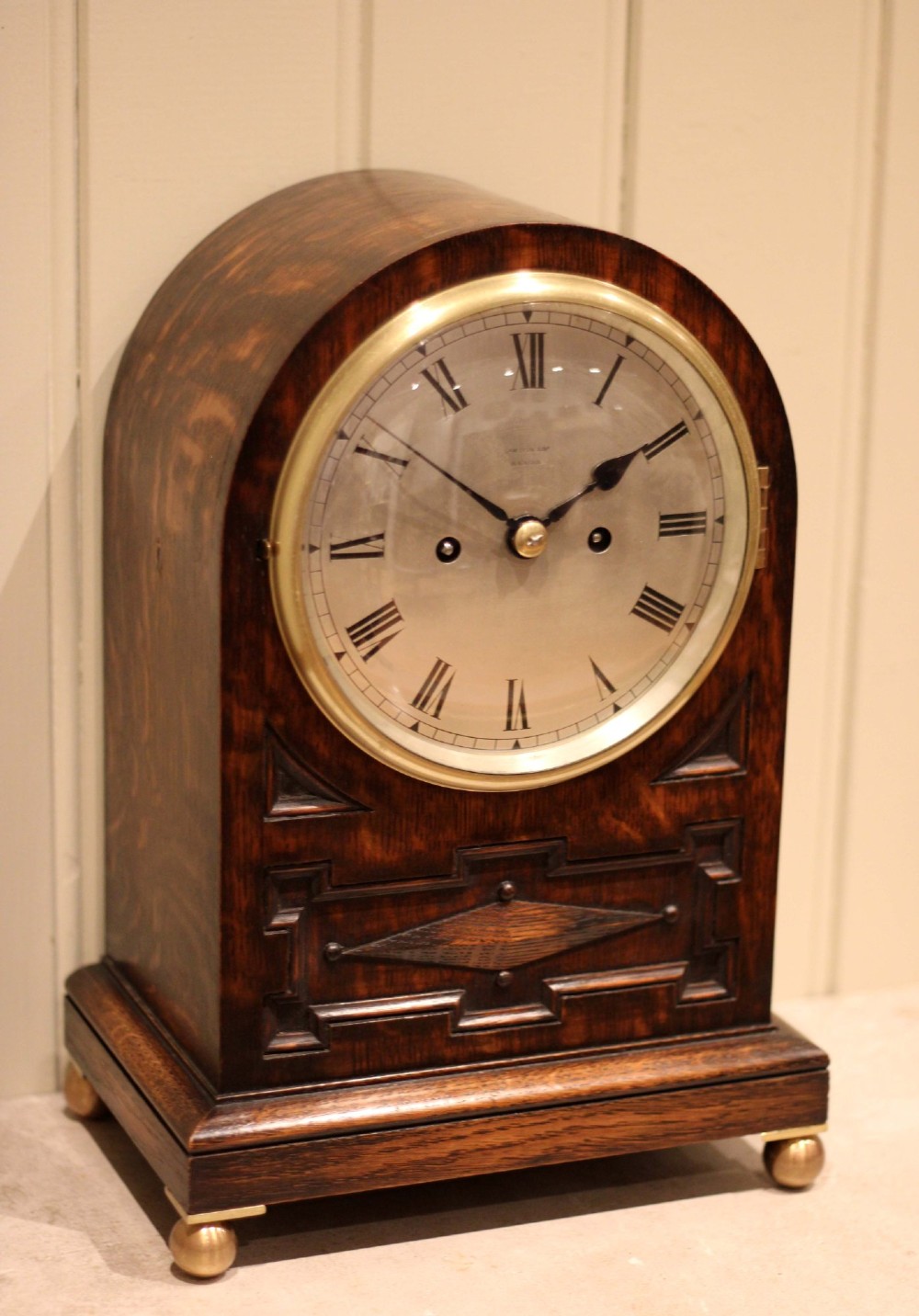 A dark oak arched-top bracket clock featuring a circular silvered dial, brass bezel, geometric wooden moulding, and brass ball feet.