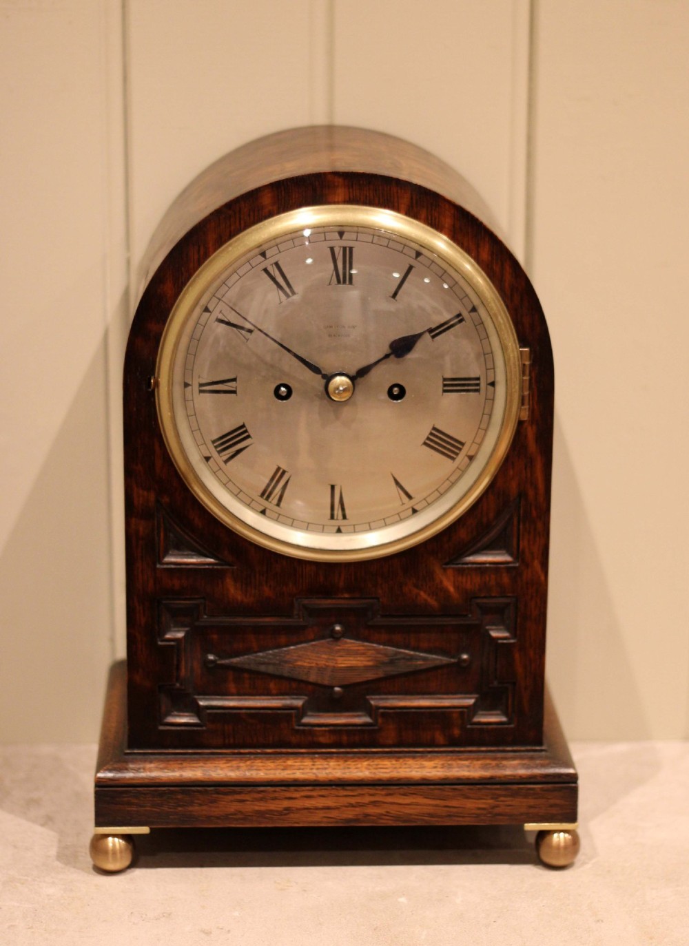 A dark oak arched-top bracket clock featuring a circular silvered dial, brass bezel, geometric wooden moulding, and brass ball feet. View II