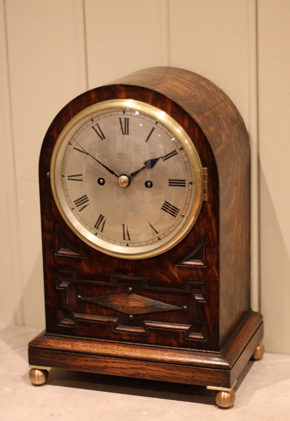 A dark oak arched-top bracket clock featuring a circular silvered dial, brass bezel, geometric wooden moulding, and brass ball feet. View III