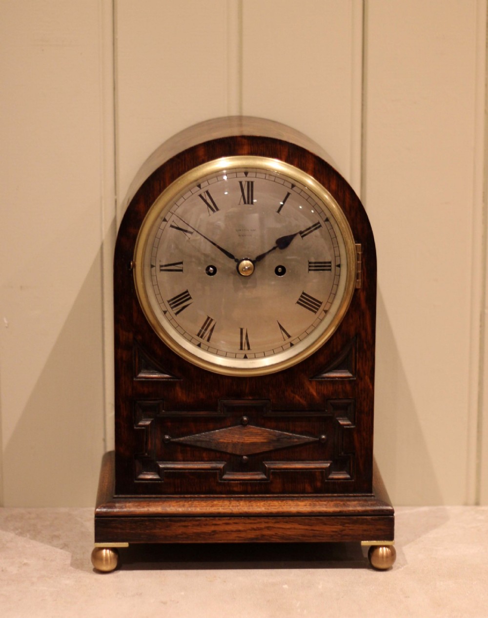A dark oak arched-top bracket clock featuring a circular silvered dial, brass bezel, geometric wooden moulding, and brass ball feet. View IV