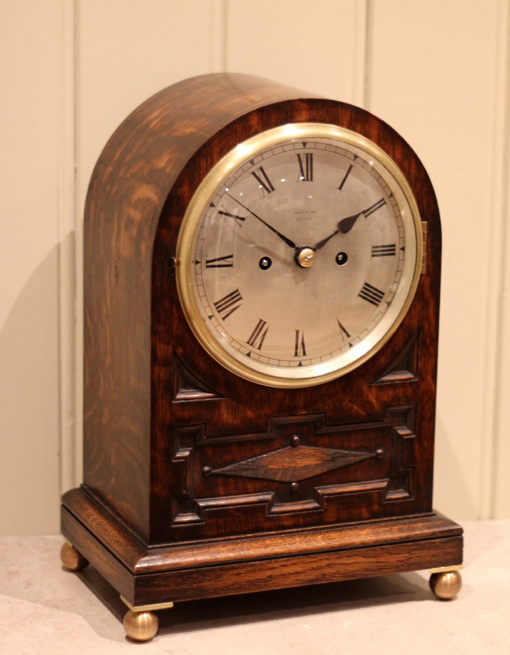 A dark oak arched-top bracket clock featuring a circular silvered dial, brass bezel, geometric wooden moulding, and brass ball feet. View V