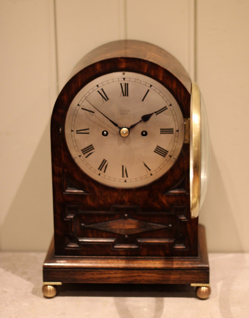 A dark oak arched-top bracket clock featuring a circular silvered dial, brass bezel, geometric wooden moulding, and brass ball feet. View VI