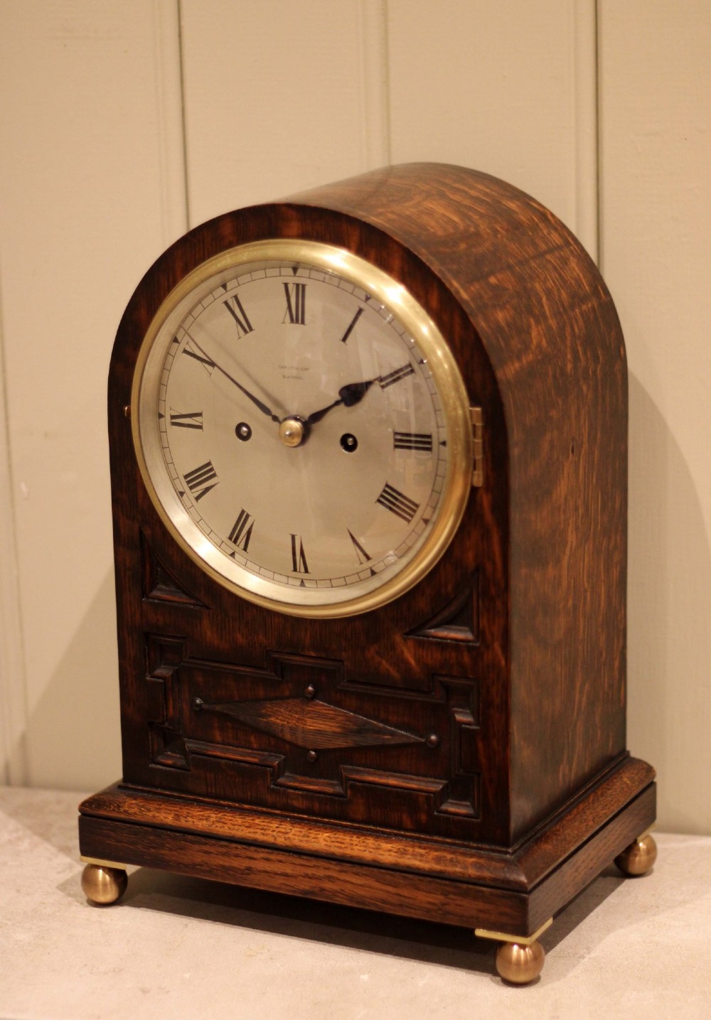 A dark oak arched-top bracket clock featuring a circular silvered dial, brass bezel, geometric wooden moulding, and brass ball feet. View VII