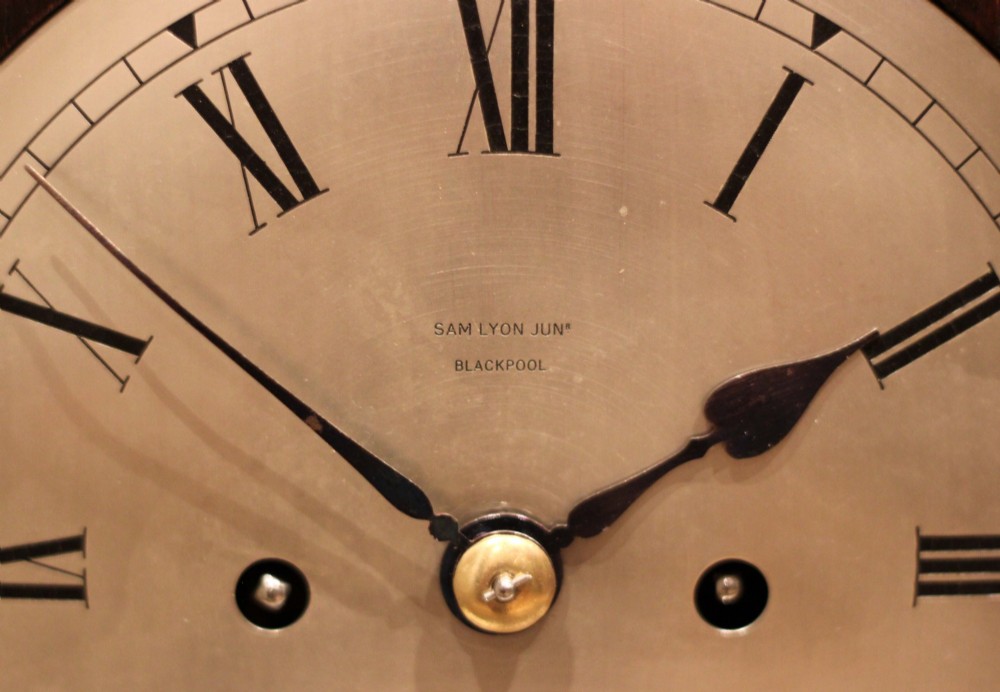 A dark oak arched-top bracket clock featuring a circular silvered dial, brass bezel, geometric wooden moulding, and brass ball feet. View VIII