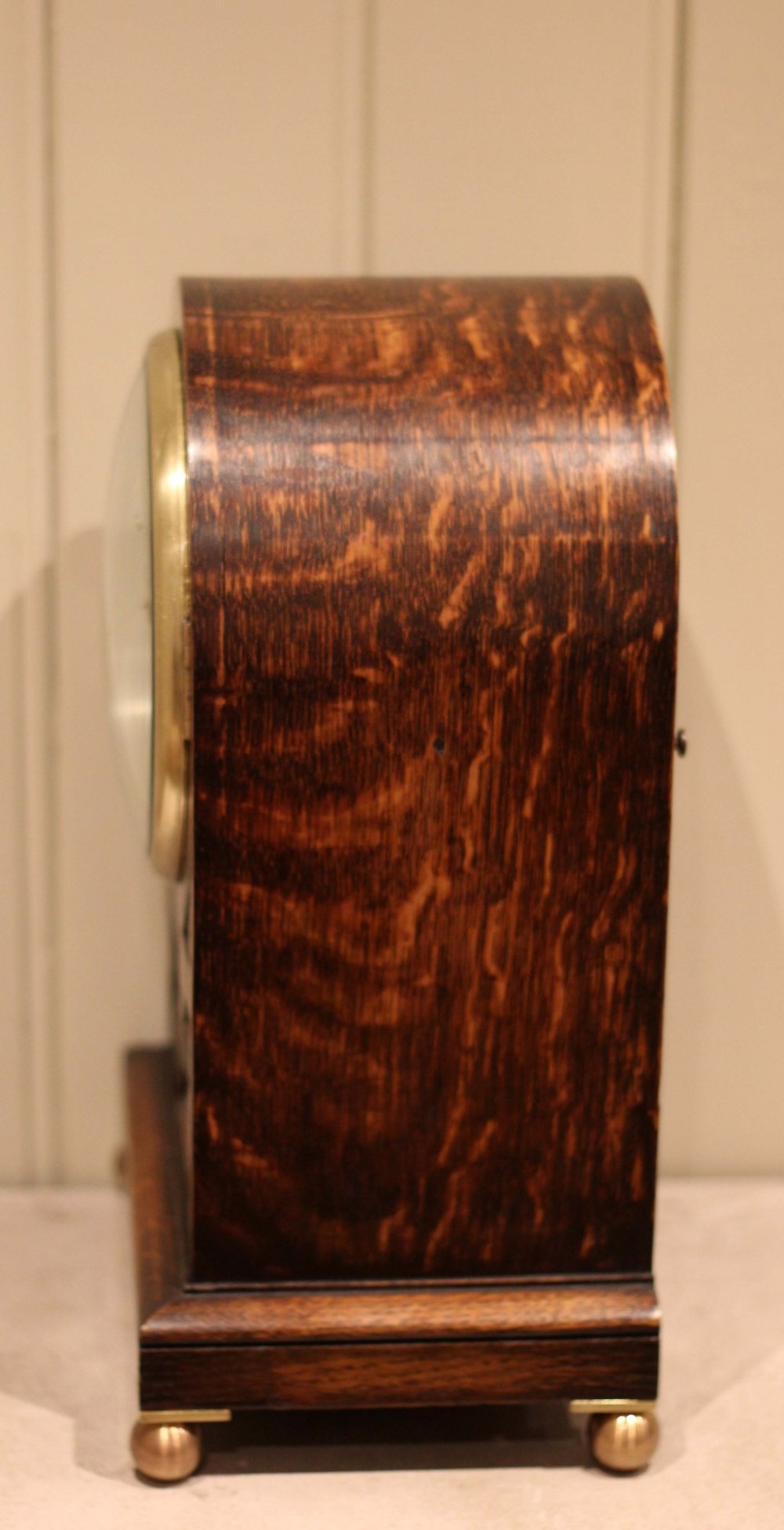 A dark oak arched-top bracket clock featuring a circular silvered dial, brass bezel, geometric wooden moulding, and brass ball feet. View IX