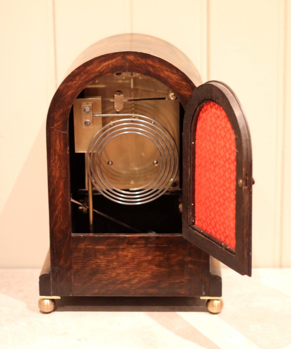 A dark oak arched-top bracket clock featuring a circular silvered dial, brass bezel, geometric wooden moulding, and brass ball feet. View XI