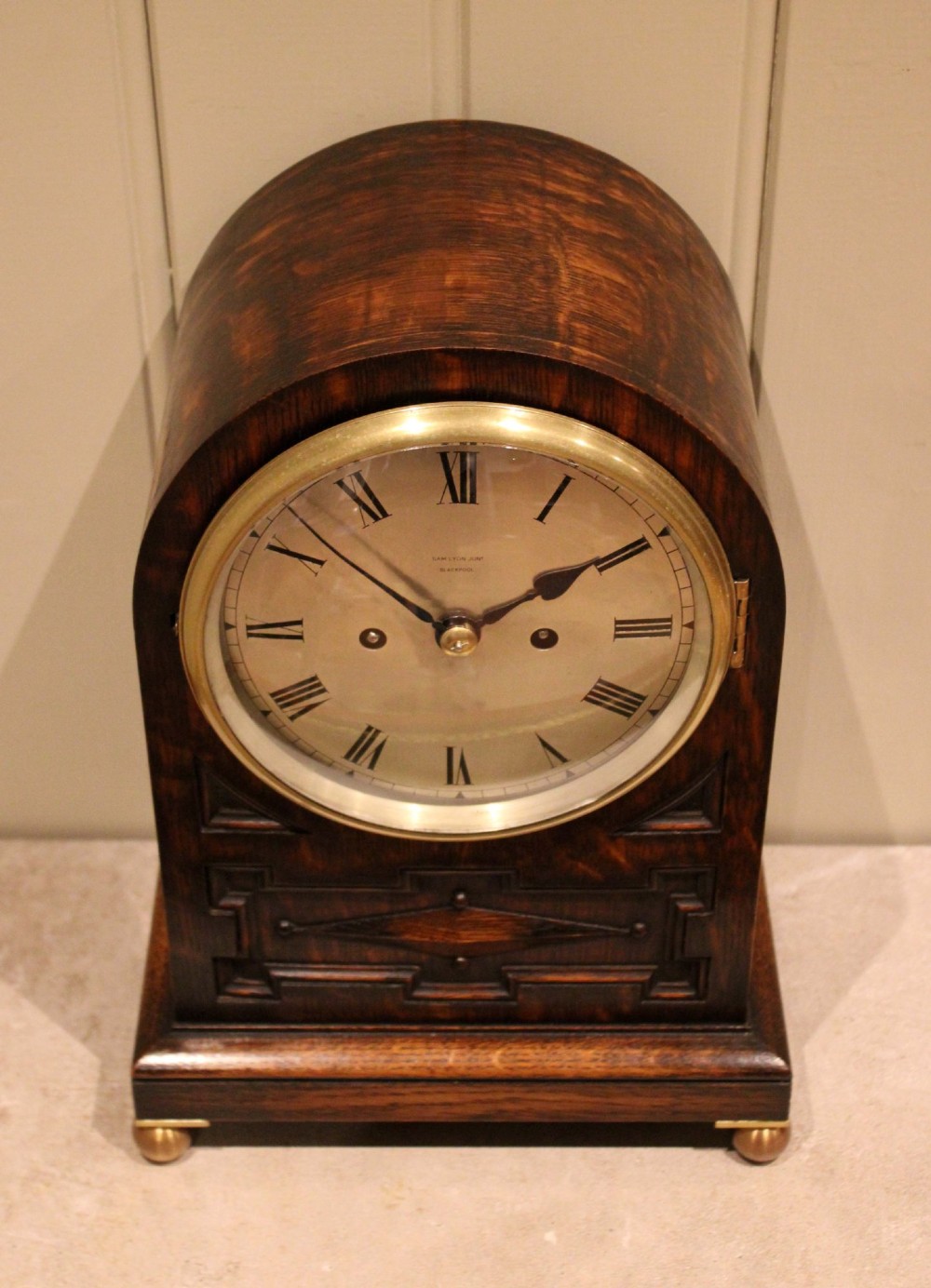 A dark oak arched-top bracket clock featuring a circular silvered dial, brass bezel, geometric wooden moulding, and brass ball feet. View XIII