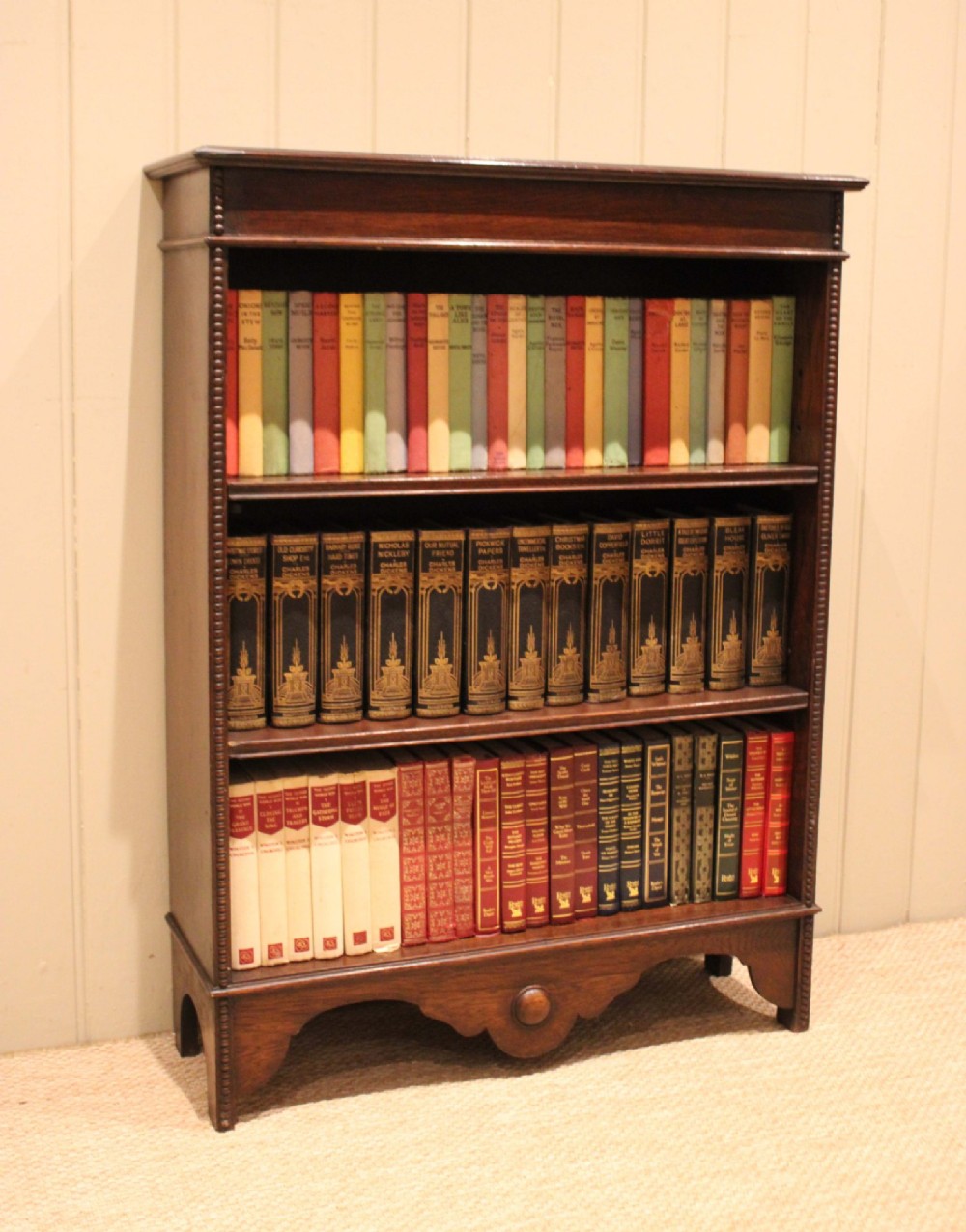 A dark oak open bookcase featuring three adjustable shelves, beaded moulding along the side columns, and a decorative shaped bottom apron with a central circular boss.