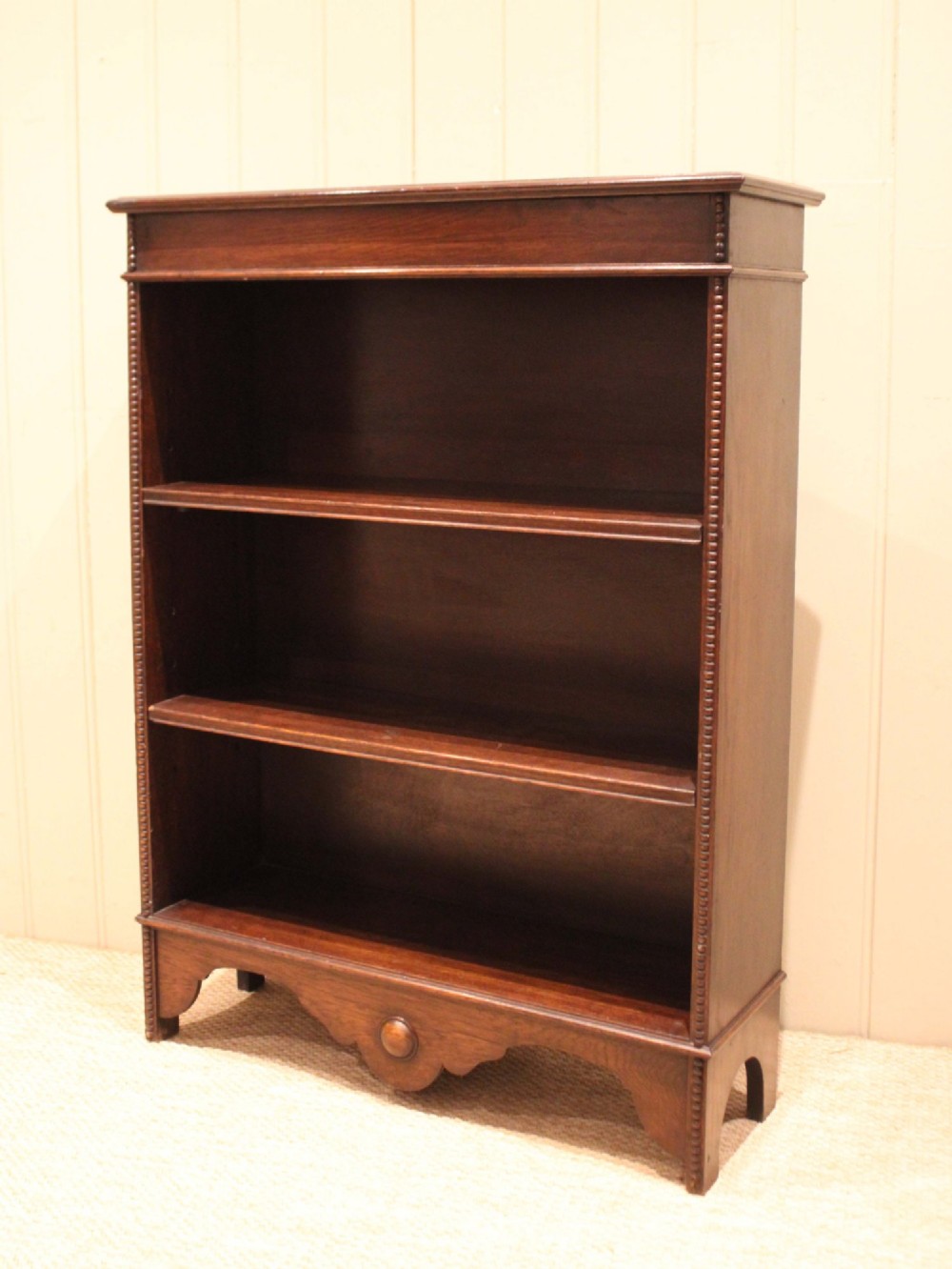 A dark oak open bookcase featuring three adjustable shelves, beaded moulding along the side columns, and a decorative shaped bottom apron with a central circular boss. View II