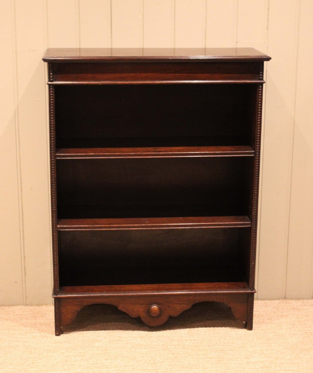A dark oak open bookcase featuring three adjustable shelves, beaded moulding along the side columns, and a decorative shaped bottom apron with a central circular boss. View VI